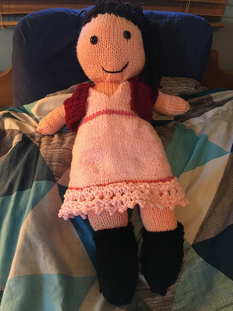 A knit doll of Aerith from Final Fantasy VII. She's got long brown hair pulled back in a twist, with bangs. She's wearing a light pink lace dress with a darker pink bands at the waist and upper hem. The hem itself is garter lace. She also has on a red jacket and black boots. Aerith from Final Fantasy VII