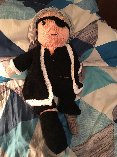 The finished doll of Akstar, with clothing. He's got long silver hair pulled back in a pony tail, and is missing his left eye, arm, and leg. He's wearing black pants and shirt, and a large black coat trimmed in white. He's got a peg leg, and the long left sleeve of his coat is tied so to stay out of his way. FFBE Akstar