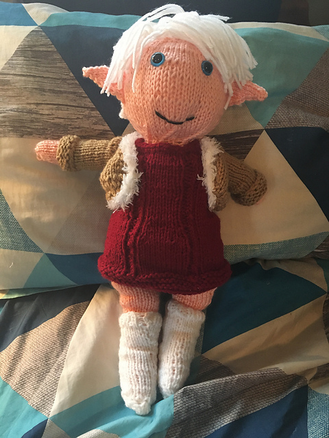 A knitted doll of Alisaie from Final Fantasy XIV. She's got a mop of white hair on her head, blue eyes, and pointy ears. She's wearing a short red dress with black shorts underneath, white boots, and a tan cropped jacket edged in white fur. Alisaie from FFXIV, front