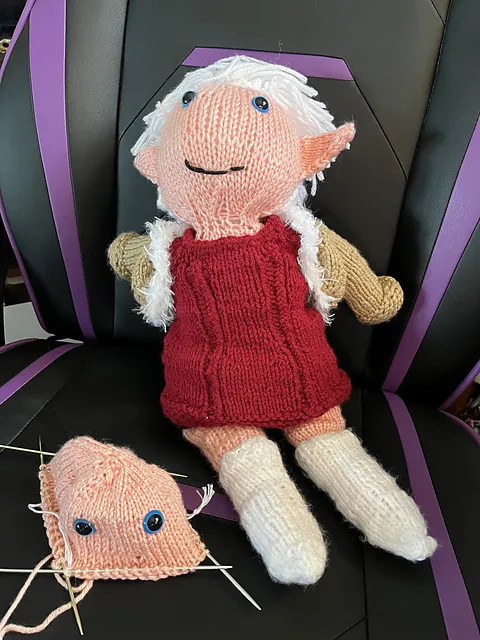 A knit doll of of Alisaie from Final Fantasy XIV, seated in a gaming chair. Next to her is the start of Alphinaud's head. It's on four double-pointed needles and has been knit long enough to place his blue eyes. Alisaie, supervising