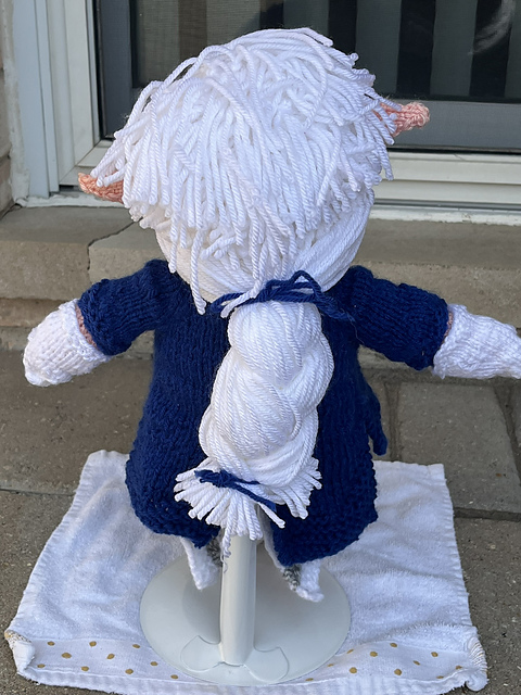 A view of the back of a knitted doll of Alphinaud from Final Fantasy XIV, in his Sage outfit from the Endwalker expansion. He's wearing a long blue coat with split tails, white boots, and has messy white hair on the top of his head and transitions into a neat braid. Alphinaud, Back