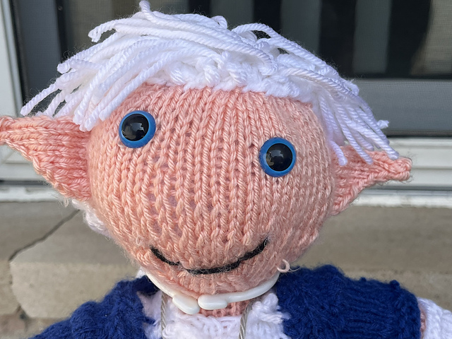 A close-up view of a knitted doll of Alphinaud from Final Fantasy XIV. His face fills the frame. He's got pointed ears, a mop of white hair, and blue eyes. Alphinaud, Close-up