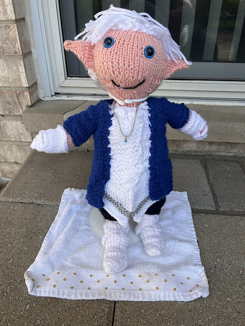 A full view of a knitted doll of Alphinaud from Final Fantasy XIV, in his Sage outfit from the Endwalker expansion. He's wearing a white tunic with pointed tails, black knee-length pants, white knee-high boots, a royal blue coat, and white gloves. Alphinaud