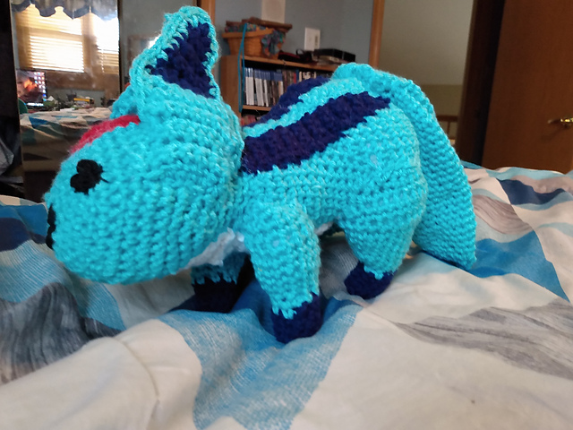 A crocheted softie of Carbuncle, a summoned creature from the Final Fantasy series. He's teal in color, with three tails and large pointed ears. He's got twin navy stripes down his back and is on all fours, with navy colored feet. A large ruby (depicted by a red crocheted patch) is on his forehead. FFXIV Carbuncle