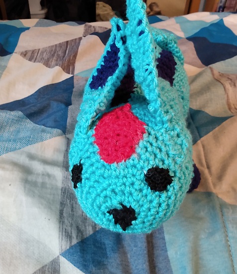 A crocheted softie of Carbuncle, a summoned creature from the Final Fantasy series. He's teal in color, with three tails and large pointed ears. He's got twin navy stripes down his back and is on all fours, with navy colored feet. A large ruby (depicted by a red crocheted patch) is on his forehead. This is a view from the top, showing off his ruby and ears. FFXIV Carbuncle, Top View