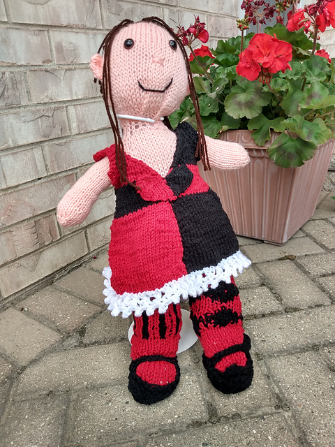 A knit doll of my Hyur from Final Fantasy XIV, wearing the Valentione Rose Attire Set. She's got brown hair pulled back in a ponytail with long bangs framing her face. She's wearing a dress, quartered in black and red with a white lace hem. The dress has a sweetheart neckline and features a red and black heart on the bodice. She's also wearing tights: the right leg is stripes of black on red, and the left leg features black diamonds on red. A pair of knitted Mary Jane style shoes completes the outfit. FFXIV Fan Fest 2023 Entry - Valentione Rose Attire Set
