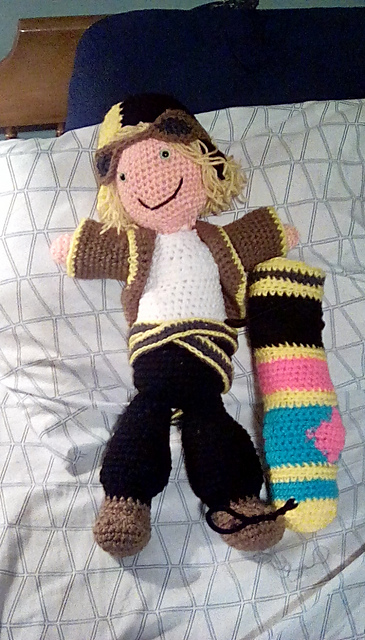 A crocheted doll of Olive, a powerful unit from Final Fantasy Brave Exvius. She's a cannoneer, wearing a leather-style jacker and goggles, with a bright yellow shoulder cannon as tall as she is by her side. FFBE's Olive