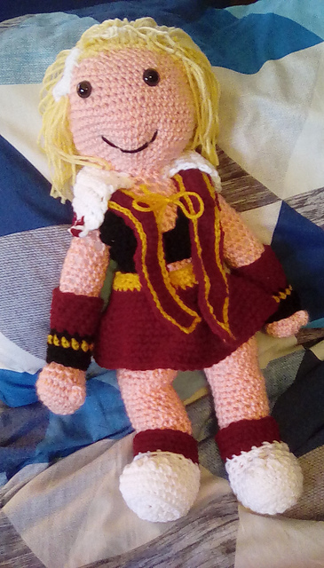 A crocheted softie of the Fina from Final Fantasy Brave Exvious, in her Season 2 outfit. She's got blond hair with a white flower in it, and is wearing a black sleevless v-neck shirt, red skirt, red scarf, and red and black gauntlets, and white shoes. Fina from FFBE
