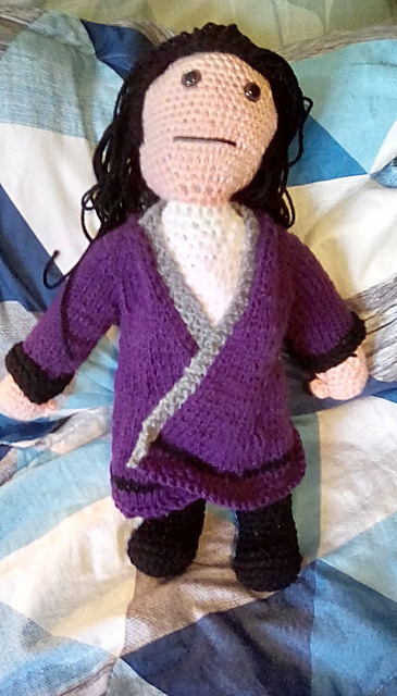 A crocheted doll of Lasswell, my favorite character from Final Fantasy Brave Exvius. He's got a white shirt and black pants on, covered by a large purple coat. He's got long black hair and a neutral expression on his face. FFBE's Lasswell