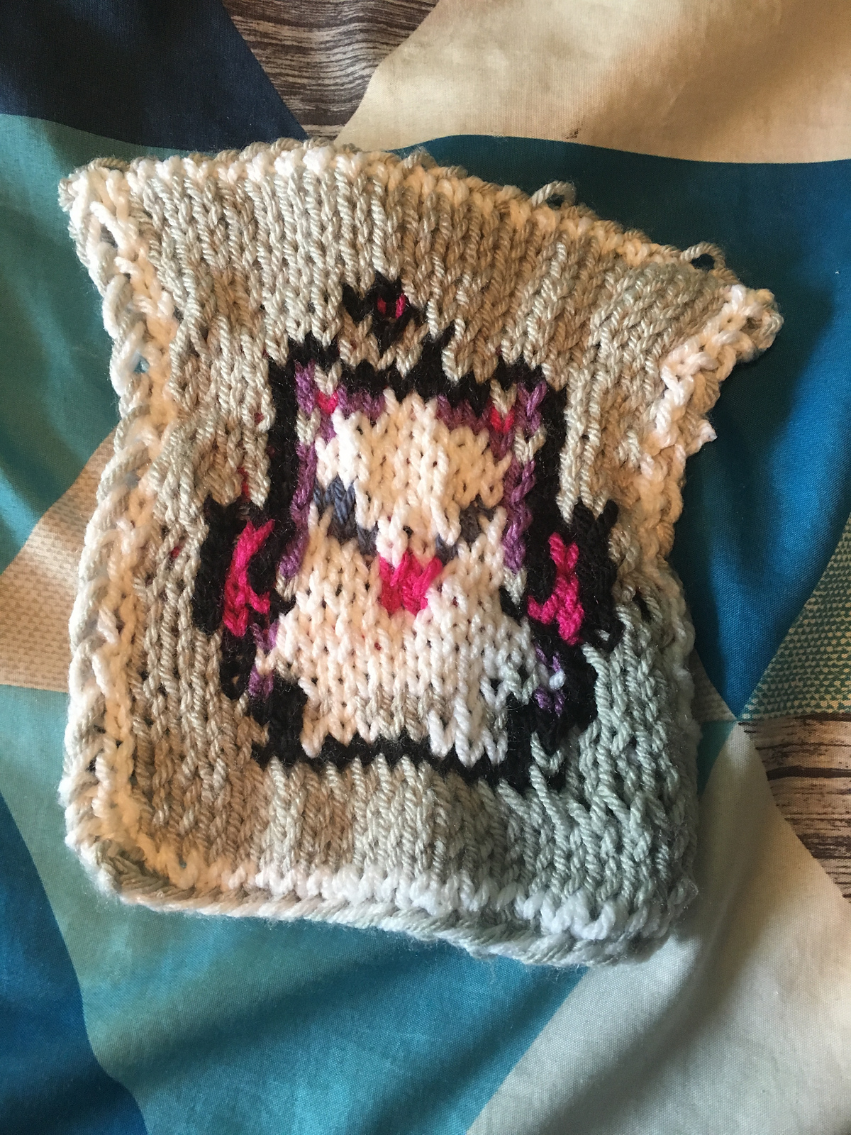 A knitted blanket square, featuring the iconic Moogle sprite from the Final Fantasy video game series. The white moogle is outlined in black against a gray background, and has pink wings and purple shadowing on its face. This is the front view. Moogle Square, front