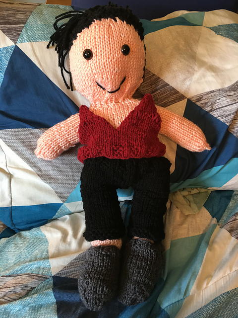 A knitted doll of Morrigan from Dragon Age: Origins. She's got black hair pulled up in a messy bun, and is wearing a sleeveless red v-neck shirt with black pants and gray boots. Morrigan from Dragon Age: Origins