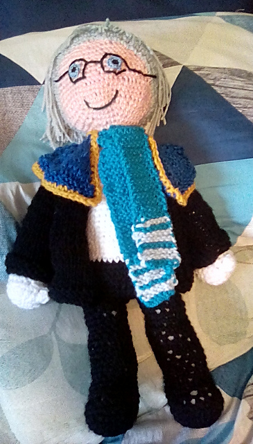 A crocheted doll of Nichol from Final Fantasy Brave Exvius, wearing knitted clothing. He's got a black jacket and pants on, with a white shirt. On his shoulders is a navy blue cape trimmed in gold. He's also wearing a teal scarf with white stripes. He's got silver hair, blue eyes, and embroidered glasses. FFBE Nichol
