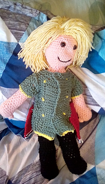 A crocheted doll of Rain, the main character from Final Fantasy Brave Exvius. He's got a shock of blond hair, and is wearing black pants, a green tunic, and a red kilt. FFBE's Rain