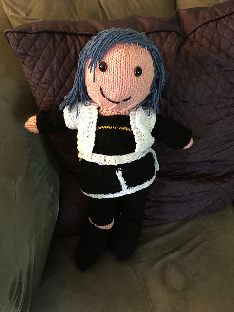 A knit doll of Maria from Star Ocean: Anamnesis.She's got blue shoulder-length hair with long bangs brushed to one side. She's wearing a black shirt with a gold stripe on it, with black leggings and a black skirt edged in white. Over this, she has on a white jacket, and a white sarong is tied around her waist. Maria from Star Ocean: Anamnesis