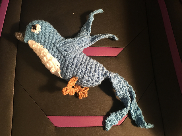 A crocheted bird softie. It's blue in color with a dark beak and white belly. It's got long pointed wings and a long split tail, so pretty much looks like two tails. Starbird, from Final Fantasy XIV