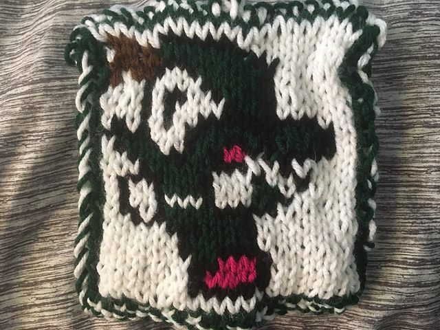 A knitted blanket square, featuring the iconic thief sprite from Final Fantasy I. The thief is outlined in black, and is wearing green clothing against a white background. He's got brown hair and pink boots. Thief Square