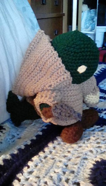 A crocheted Tonberry from the Final Fantasy series. It is green in color with yellow eyes. It's wearing a tan robe and carrying a lantern in its left hand and a knife in its right. This view is from the side, so you can see the hood on the robe. Tonberry 1