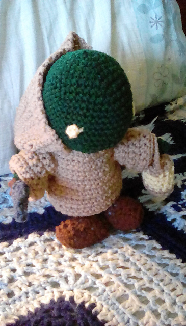 A crocheted Tonberry from the Final Fantasy series. It is green in color with yellow eyes. It's wearing a tan robe and carrying a lantern in its left hand and a knife in its right. This view is from the front. Tonberry 2