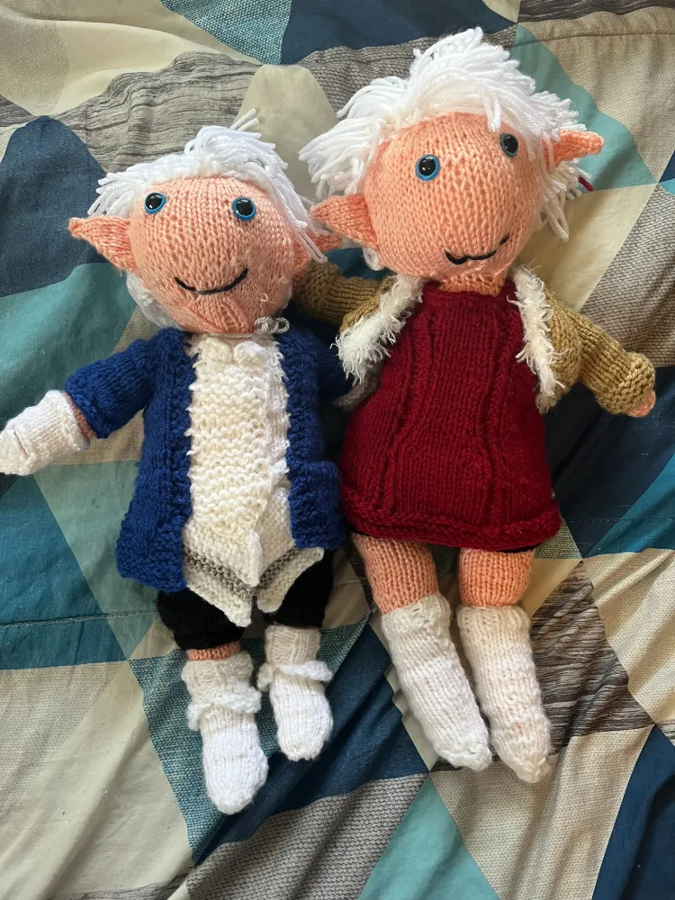 Knitted dolls of Alphinaud (left) and Alisaie (right) from Final Fantasy XIV. Both have pointed ears, white hair, and blue eyes. Alphi is in blue and white, and Alisaie is in red, white, and tan. Alisaie is bigger than her twin. Twins Together