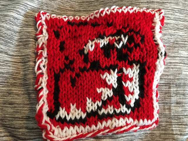 A knitted blanket square, featuring the iconic White Mage sprite from the Final Fantasy video game series. The Mage is outlined in black, wearing a red hoode robe with white at the hems, and is on a red background. White hair peeks out from under the hood. This is the back view. White Mage Square