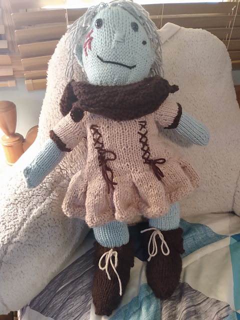 A knitted doll of one of my FFXIV characters, Windy Storm. She's got light green skin and silvery-gray short hair. She's wearing the Wayfarer's Tunic set: a light brown tunic, pleated, with lacing at the princess seams. Tied around her shoulders is a long brown cape with a large hood. Brown boots with light brown laces complete the outfit. Windy Storm, a Warrior of Light from FFXIV, front